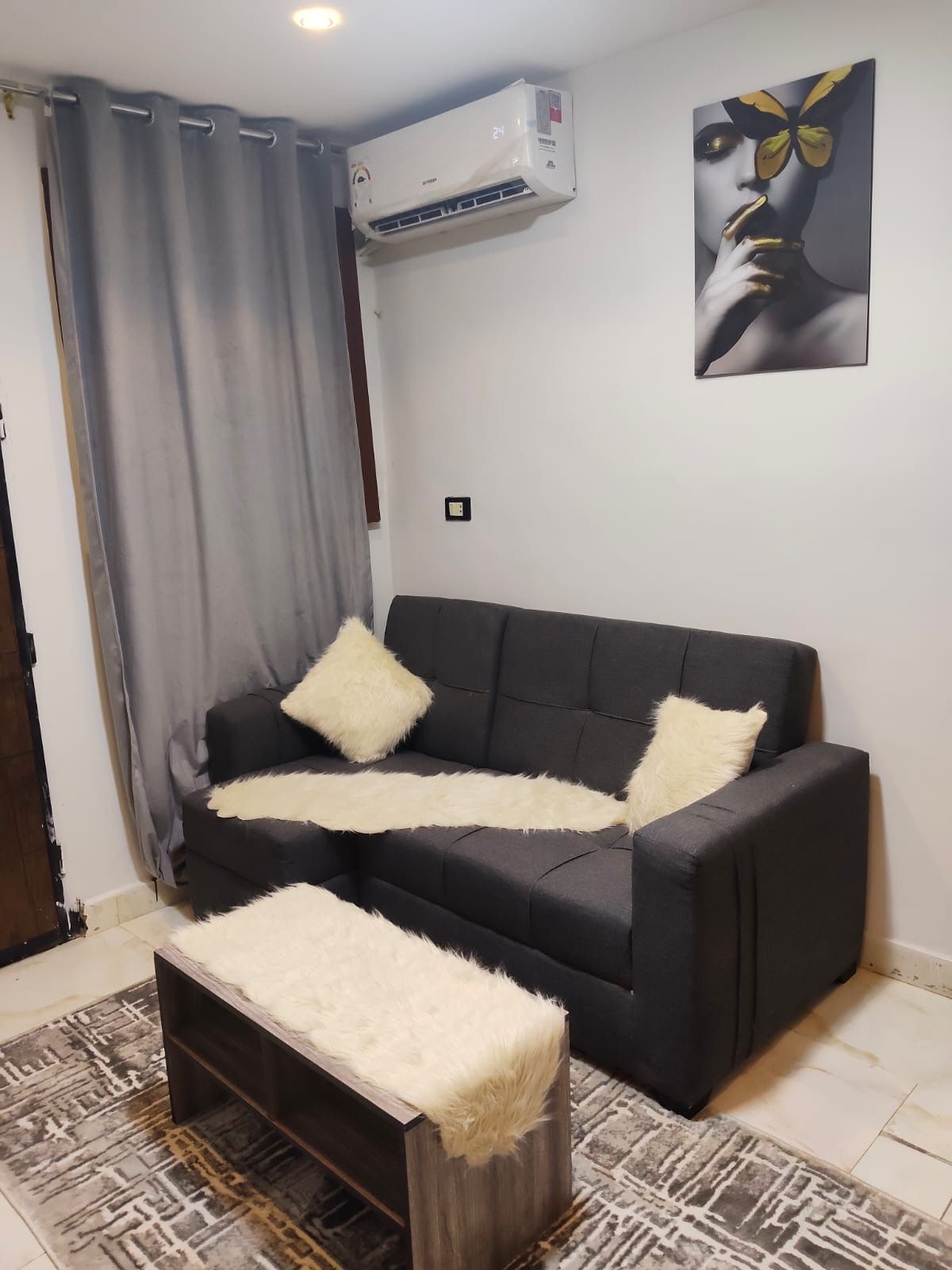 A living room with a black sofa, two white fluffy cushions, a wooden bench with a white fluffy cover, a patterned rug, a wall-mounted air conditioner, and a framed artwork.