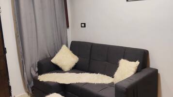 A living room with a black sofa, two white fluffy cushions, a wooden bench with a white fluffy cover, a patterned rug, a wall-mounted air conditioner, and a framed artwork.