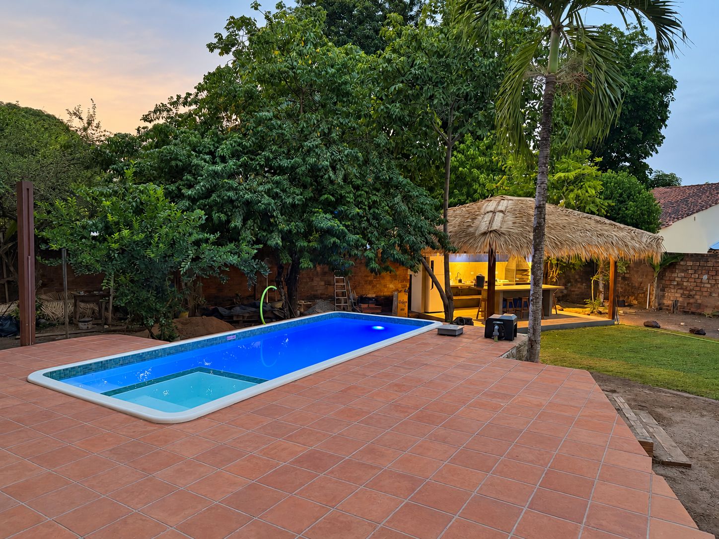 Una piscina con borde de baldosas, rodeada de árboles y una estructura con techo de paja.