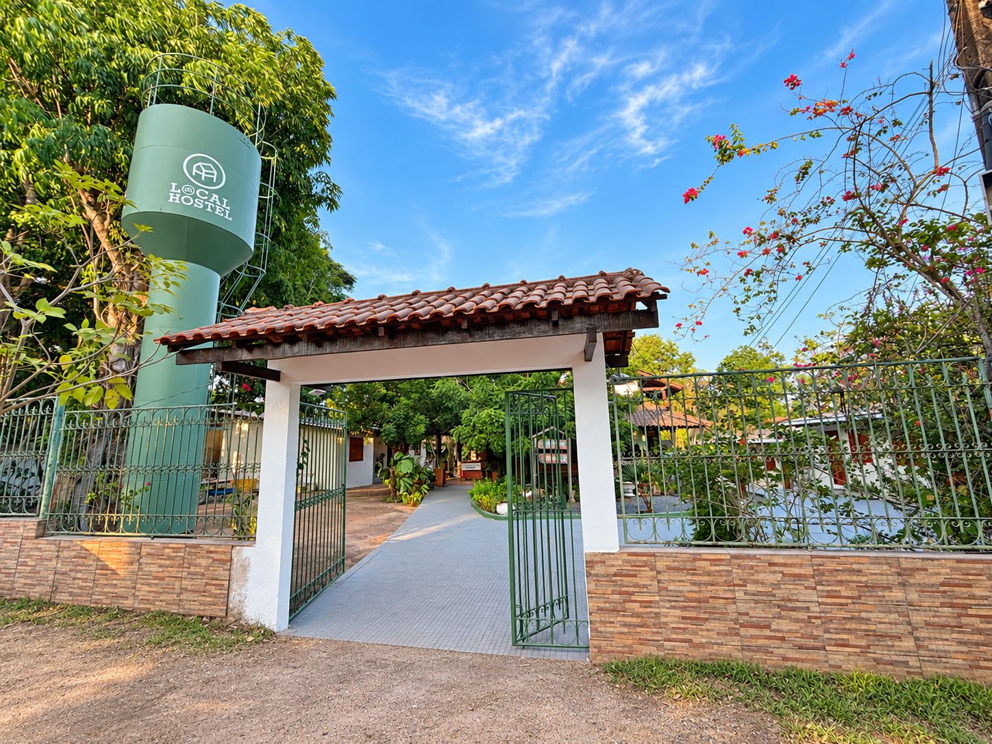 Entrada de un hostal con un corredor cubierto, rejas metálicas y un tanque cilíndrico verde.