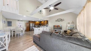 Ein geräumiger Wohnbereich mit grauem Sofa, einem Esstisch mit Stühlen, einer Küche mit Holzschränken und einem Deckenventilator.