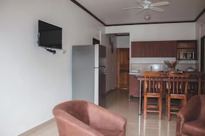 A living room with a flat-screen TV mounted on the wall, a dining area with a wooden table and chairs, and a kitchen with wooden cabinets and a refrigerator.
