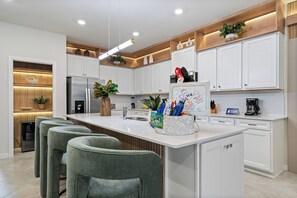 A modern kitchen with white cabinetry, a central island with bar stools, and a built-in oven. - Brand New Disney-Themed 8BR Home | Windsor Cay (Clermont)