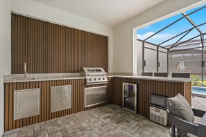 Eine Außenküche mit eingebautem Herd, Backofen und Kühlschrank, einem Boden aus Ziegelsteinen und Blick auf den Pool.