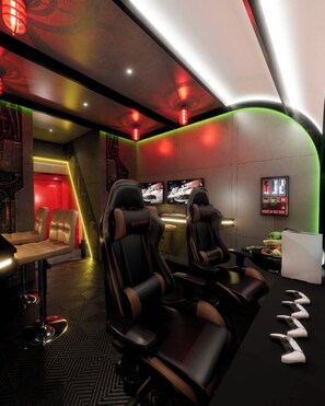 A gaming lounge with two gaming chairs, a table with a chair, and a wall-mounted screen. - Brand New Disney Themed 10BR Home Windsor Cay Res (Clermont)