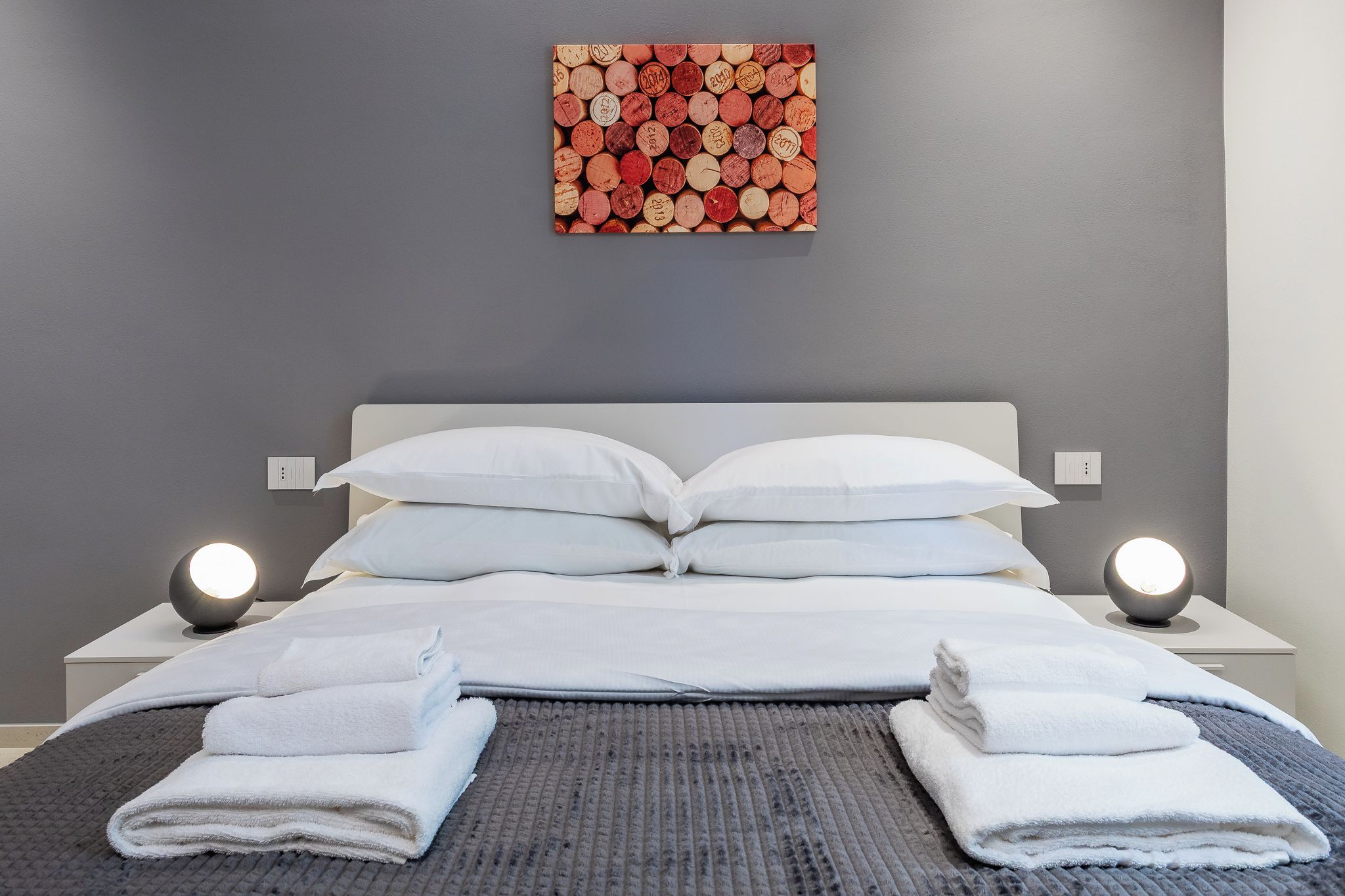 A neatly made bed with white linens and towels, a grey textured bedspread, and a wall with a corkboard.