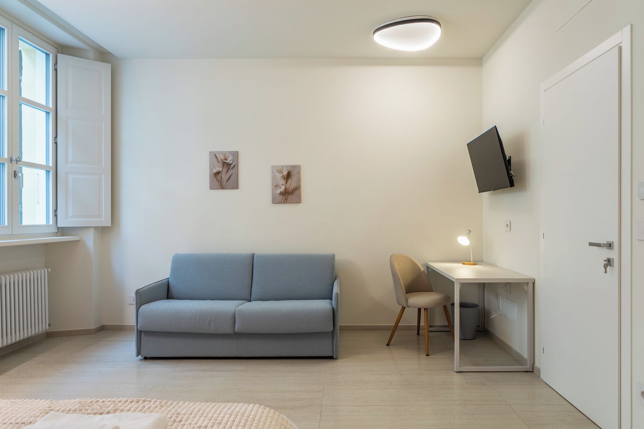 A modern living room with a light blue sofa, a small desk with a chair, and a flat-screen TV mounted on the wall.