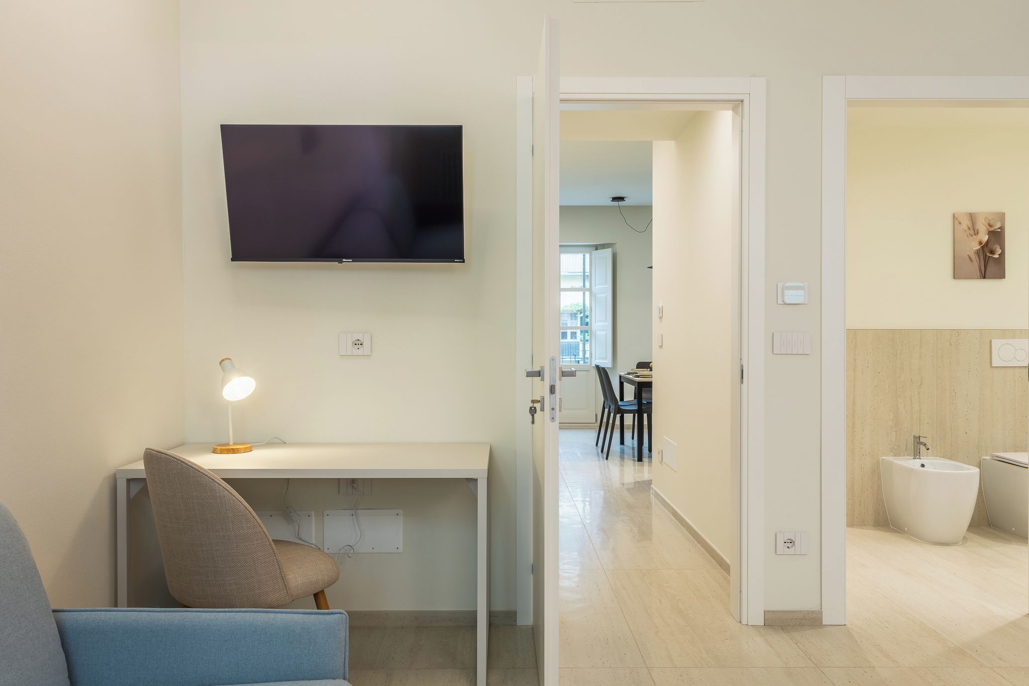 Un soggiorno moderno con una TV a schermo piatto, una scrivania con una lampada e una sedia. Un corridoio conduce a una sala da pranzo e un bagno con bidet.