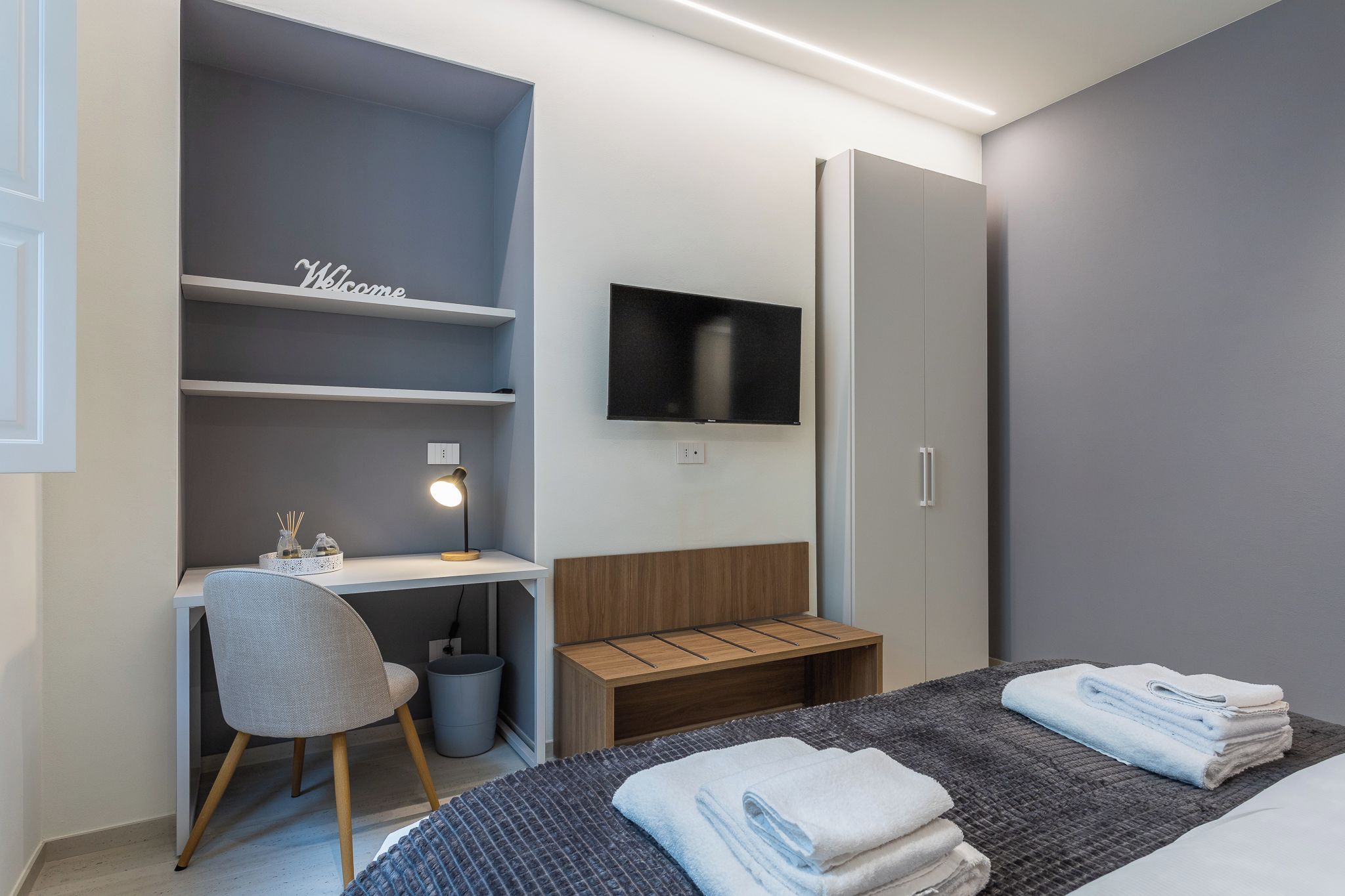 Une chambre d’hôtel moderne équipée d’un bureau, d’une chaise, d’une télévision, d’une armoire et de serviettes soigneusement pliées.