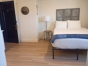 A bedroom with a bed, a bedside table, a lamp, a clock, and a door. - 310 - Historic Charm at The George - New 1BR (Klamath Falls)