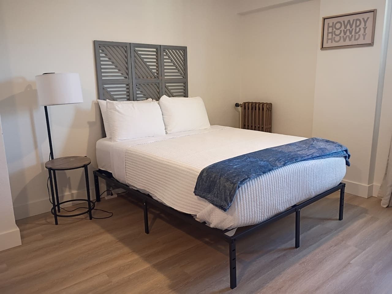 A bedroom with a bed, a side table, a lamp, a radiator, and a wall-mounted sign.