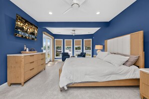 Quarto com cama grande, penteadeira de madeira, televisão na parede e vista para o exterior pelas janelas.