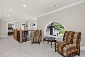Um quarto espaçoso com teto alto, uma janela grande com vista para o exterior, escada de madeira e um espaço de estar com poltrona estampada e uma mesa pequena.