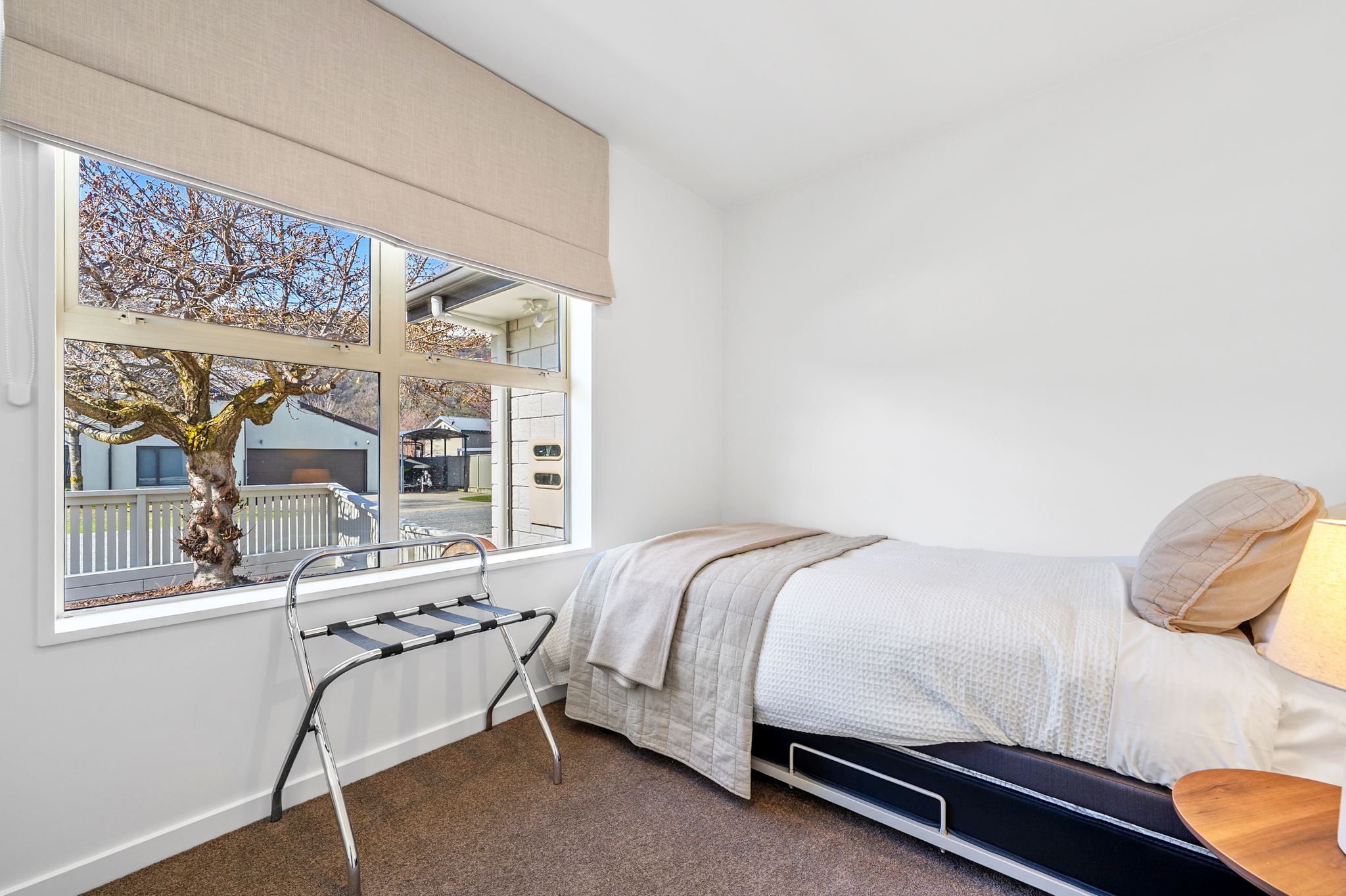 A bedroom with a bed, a window with a view of a tree and buildings, a radiator, and a bedside table with a lamp.