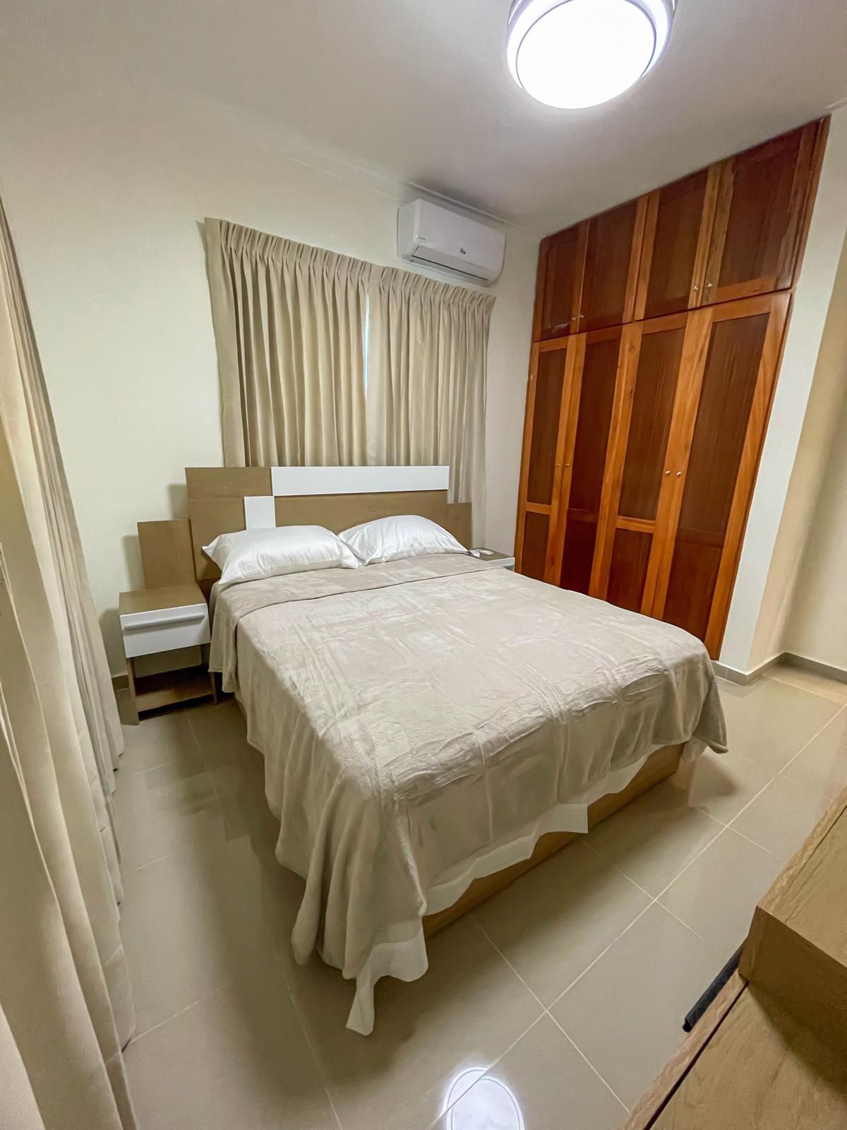 A bedroom with a bed, nightstand, wardrobe, and an air conditioning unit.