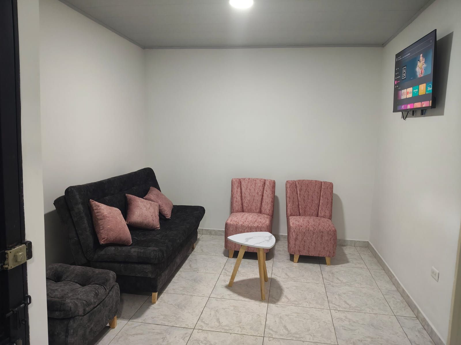 A living room with a black sofa, two pink armchairs, a small white table, and a TV mounted on the wall.