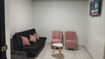 A living room with a black sofa, two pink armchairs, a small white table, and a TV mounted on the wall.