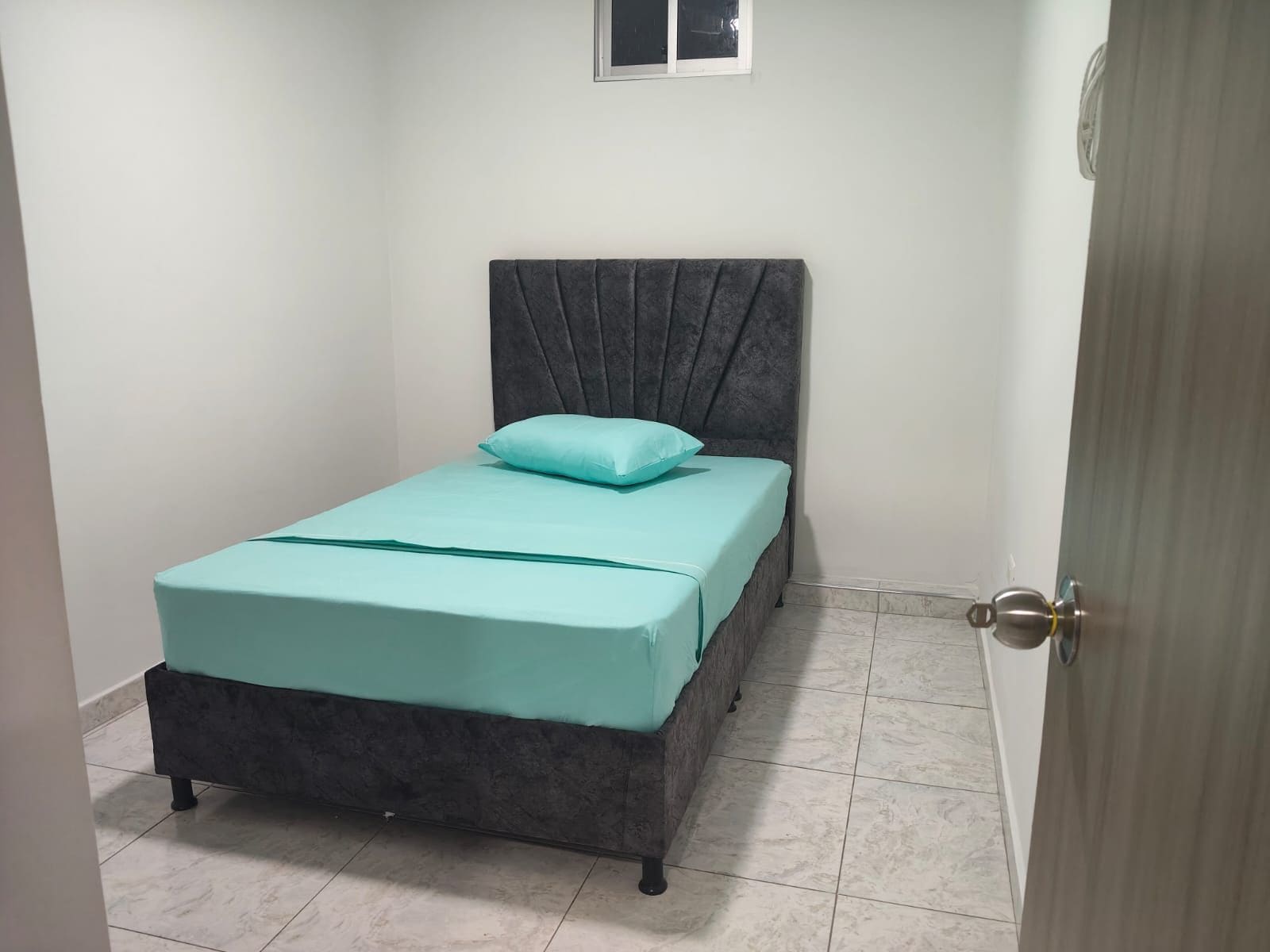 Une petite chambre avec un lit simple, dotée d’une tête de lit grise, d’une couvre-lit turquoise et d’un seul oreiller bleu clair.