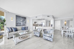 Una sala moderna con sofá, sillones y una mesa de centro. Se observa una cocina al fondo con zona de comedor.