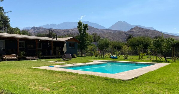A pool with a mountain backdrop, a wooden building, and a grassy lawn.