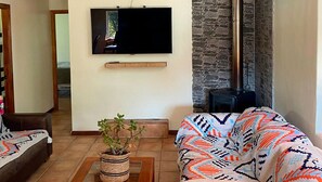 A living room with a flat-screen TV mounted on the wall, a stone fireplace, a sofa with a patterned throw, and a potted plant. - Adorable 3-bedroom 8 pax cottage in charming Vicuña, Elqui Valley awaits you (Coquimbo)