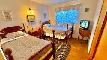 Quarto com duas camas de madeira, uma escrivaninha, uma cadeira, televisão e janela com cortinas estampadas.