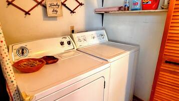 A laundry room with a front-loading washer and dryer, a shelf with cleaning supplies, and a wall-mounted rack with a sign.