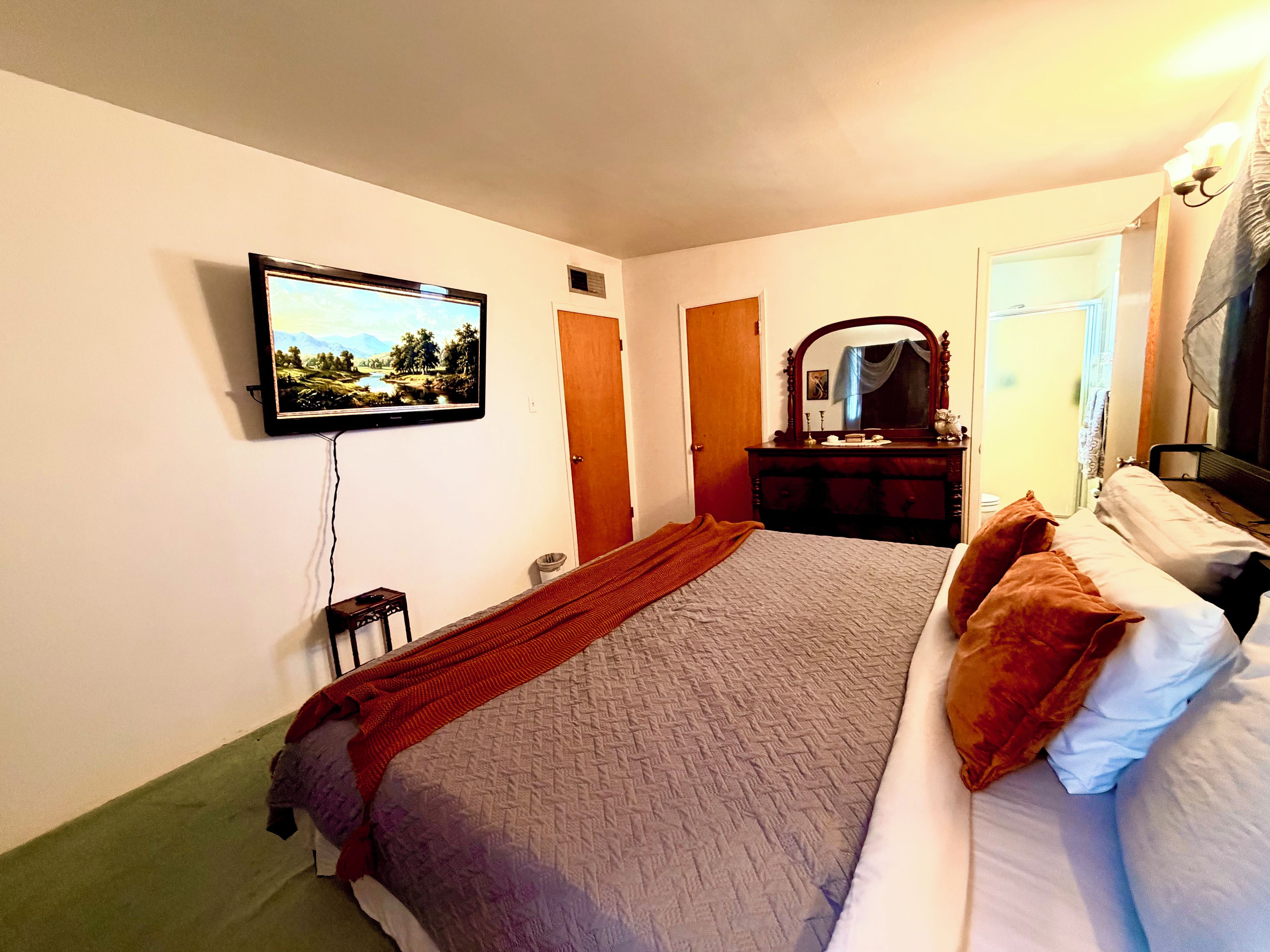 Habitación de hotel con cama, cómoda, espejo, televisor montado en la pared y puerta que da a otra habitación.