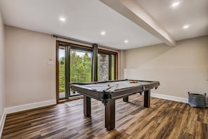 A pool table in a room with wooden flooring, a stone pillar, and a view of greenery through large windows. - Luxury Chalet in Sainte-Béatrix / Spa & Sauna / Sleeps Up to 11 People, 1 Hour from Montreal (Sainte-Bã©Atrix)