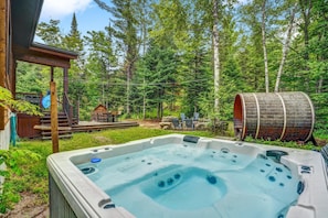 A hot tub surrounded by a wooden deck, a rustic cabin, and a forested area. - Luxury Chalet in Sainte-Béatrix / Spa & Sauna / Sleeps Up to 11 People, 1 Hour from Montreal (Sainte-Bã©Atrix)