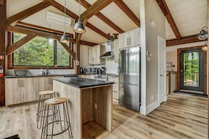 A kitchen with wooden cabinets, a stainless steel refrigerator, a large island with stools, and a view of the outdoors through the windows. - Luxury Chalet in Sainte-Béatrix / Spa & Sauna / Sleeps Up to 11 People, 1 Hour from Montreal (Sainte-Bã©Atrix)