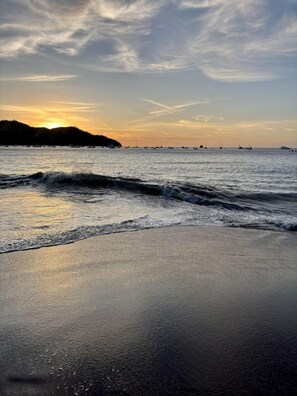 Una playa con olas, un atardecer y barcos a lo lejos.