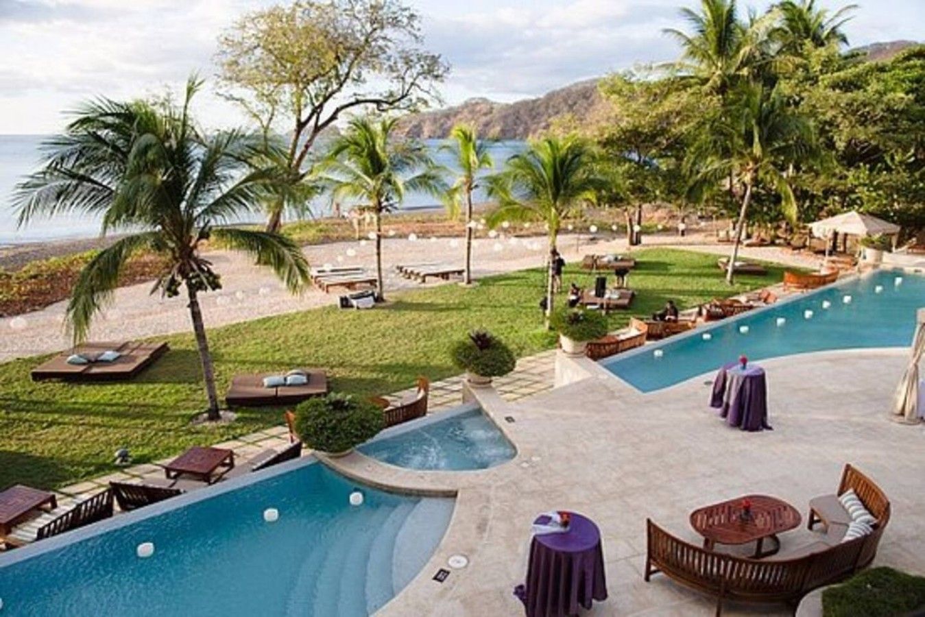 A pool area with lounge chairs, a hot tub, and a view of the sea.