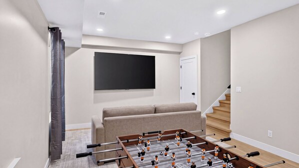 A modern living room with a foosball table, a flat-screen TV mounted on the wall, and a staircase leading to another level. - Lobo Silver Summit by Avantstay Mountain-view Balcony Game Room + Near Deer Valley (Park City)