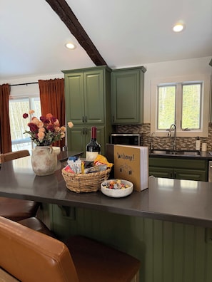 A kitchen with green cabinets, a welcome sign, a bottle of wine, and a basket of snacks. - Stoneline Chalet - Hot Tub, Sauna, Fire Pit, Game Rm (Jewett)