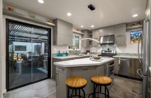 A modern kitchen with a large island, stainless steel appliances, and a glass door leading to an outdoor area. - Sunlit Oasis: Walk to Bay & Beach - Hot Tub, Fire Pit, BBQ & Wraparound Deck (San Diego)