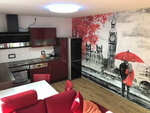 Une cuisine compacte dotée d’appareils électroménagers modernes, un coin repas avec des meubles rouges et une grande fresque murale représentant un couple sous un parapluie.