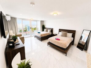 A hotel room with two beds, a desk with a lamp, a TV, and a balcony with a city view. - Elegant Beach Escape with Ocean Views Isla Verde (Carolina)