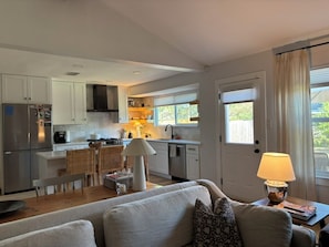 Cozinha compacta com eletrodomésticos modernos, área de jantar com mesa e cadeiras e um espaço de estar aconchegante com sofá e luminária.