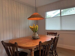 Een eetgedeelte met een ronde houten tafel, zes houten stoelen, een hanglamp en een raam met blinds.