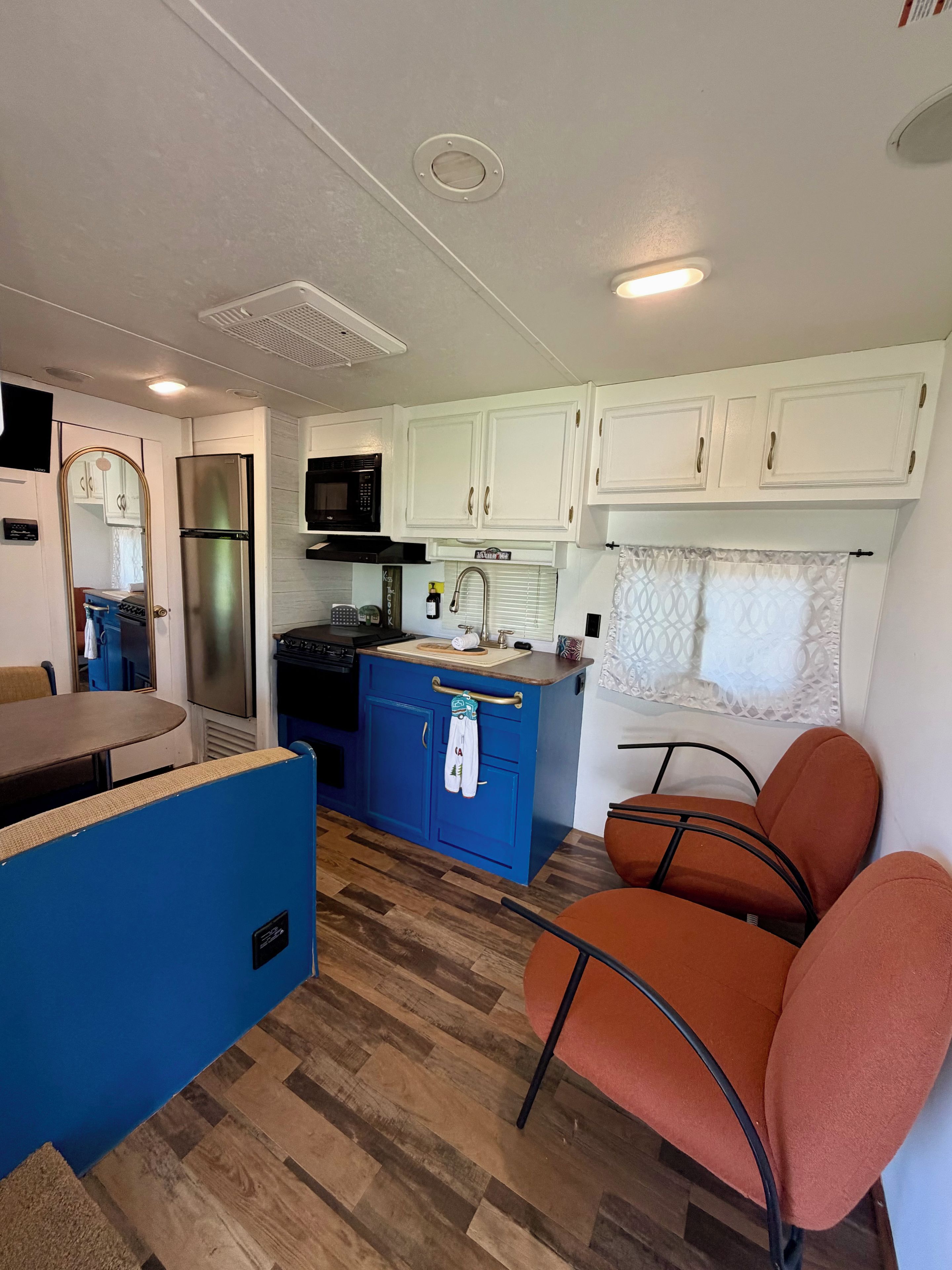 Cocina compacta con microondas, estufa y refrigerador. Hay una encimera azul y una zona de comedor con sillas.