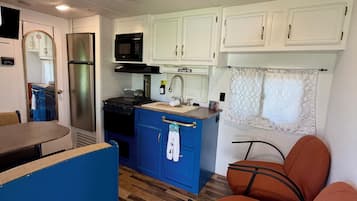 Cocina compacta con microondas, estufa y refrigerador. Hay una encimera azul y una zona de comedor con sillas.