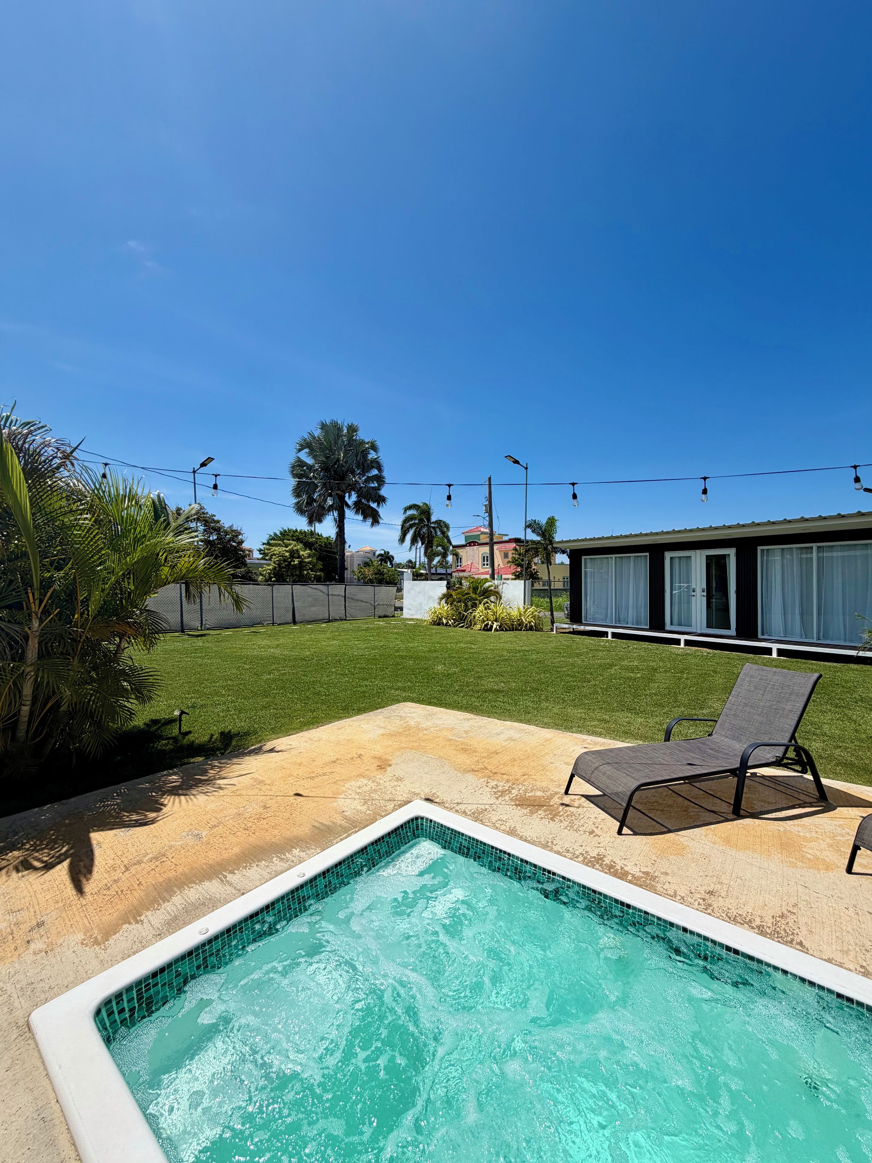 A swimming pool with a tiled edge, surrounded by a sandy area and a grassy lawn. There are two lounge chairs and a palm tree in the foreground.