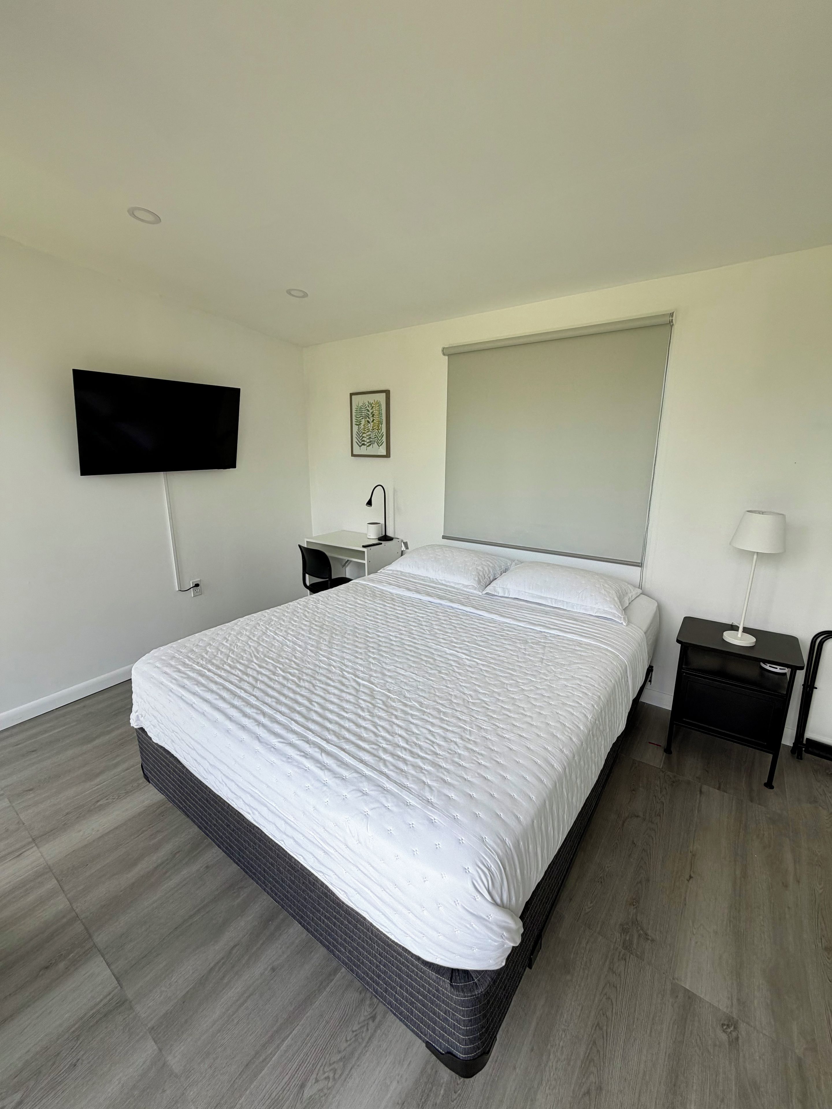 Un dormitorio moderno con una cama grande, un televisor de pantalla plana montado en la pared, una mesita de noche con una lámpara y una ventana con persiana.