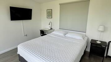 Un dormitorio moderno con una cama grande, un televisor de pantalla plana montado en la pared, una mesita de noche con una lámpara y una ventana con persiana.