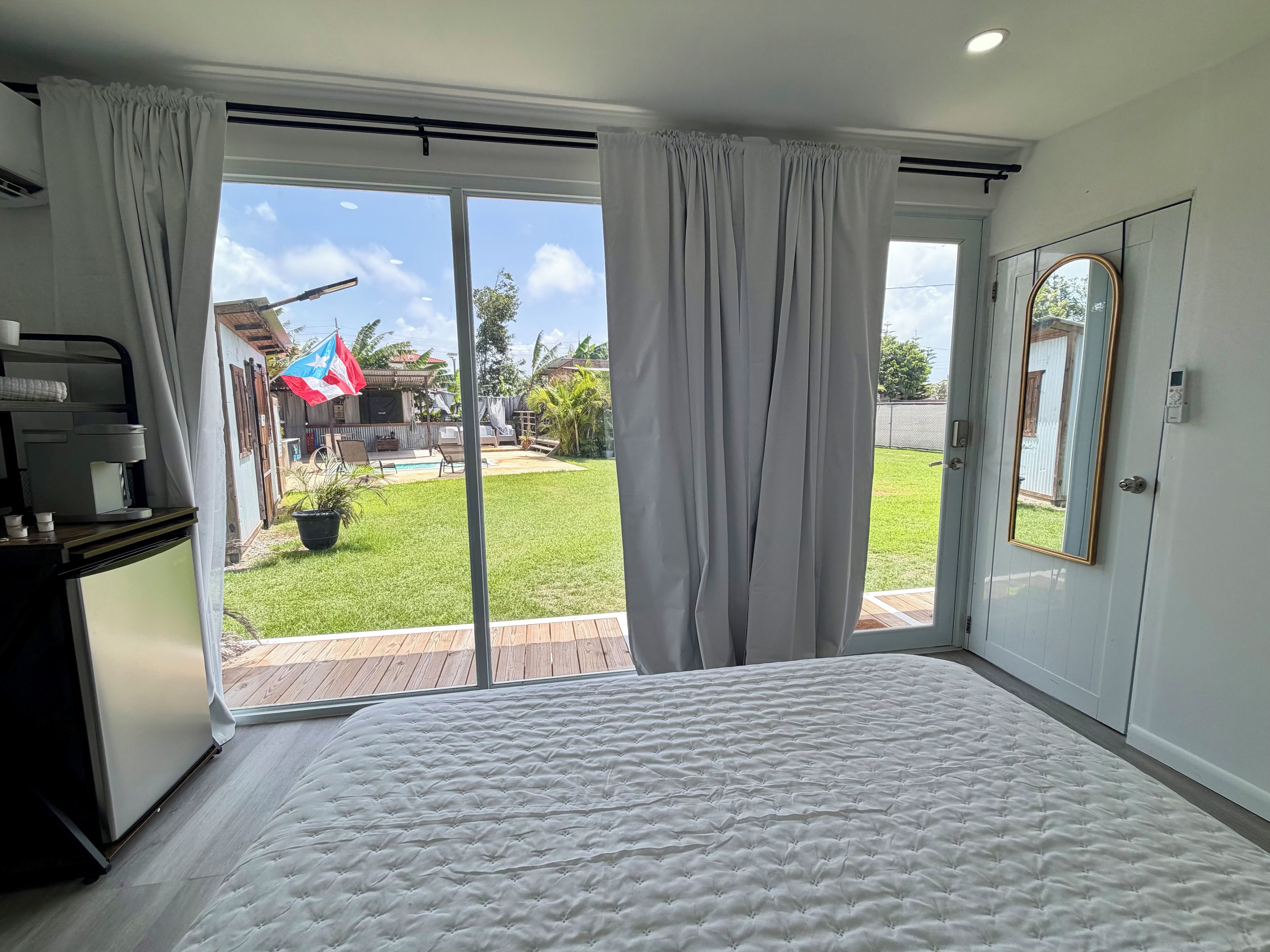 A bedroom with a large bed, sliding glass doors to a backyard, a small table with a coffee maker, and a mirror on the wall.