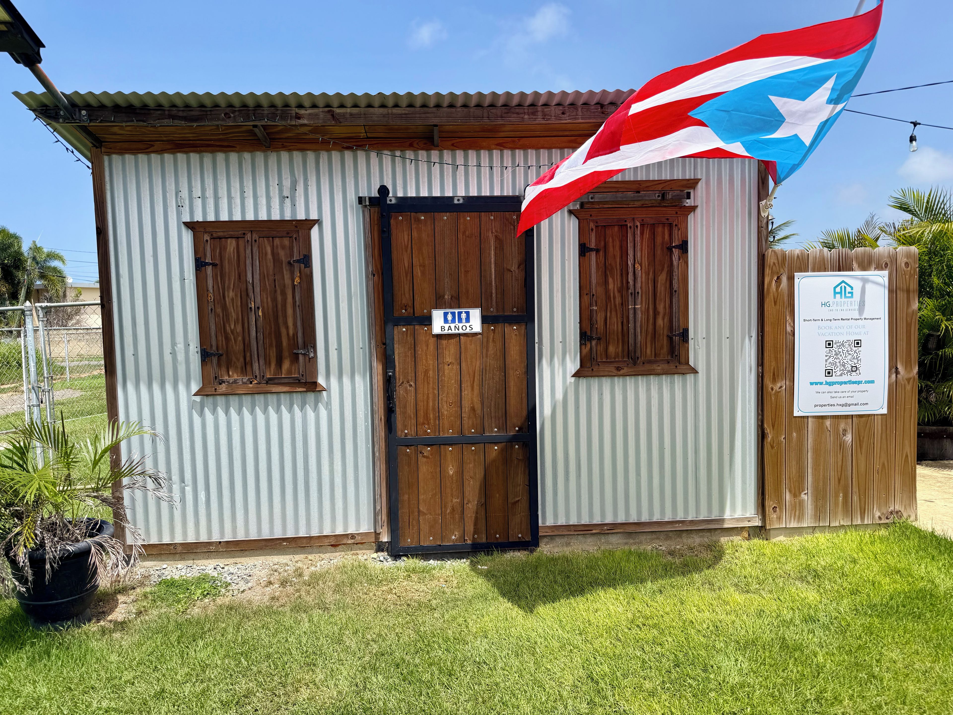 Un edificio pequeño con fachada de metal corrugado, una puerta de madera y dos ventanas con persianas. Una bandera ondea en el lado derecho.