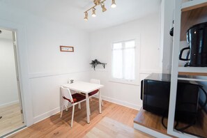 A room with a white table and chairs, a window with curtains, a wall-mounted shelf with a plant, and a coffee machine on a counter. - 2336B - Lovely Backyard Studio near UC Berkeley (Berkeley)