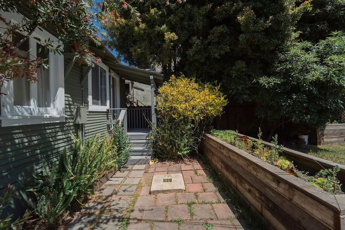 A residential property with a paved pathway leading to a front door, surrounded by greenery and a wooden fence.