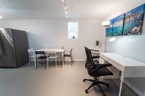 Un espacio de oficina moderno con un escritorio blanco, una silla de oficina negra y una mesa de comedor con sillas. Hay un refrigerador y una ventana con vista al mar.
